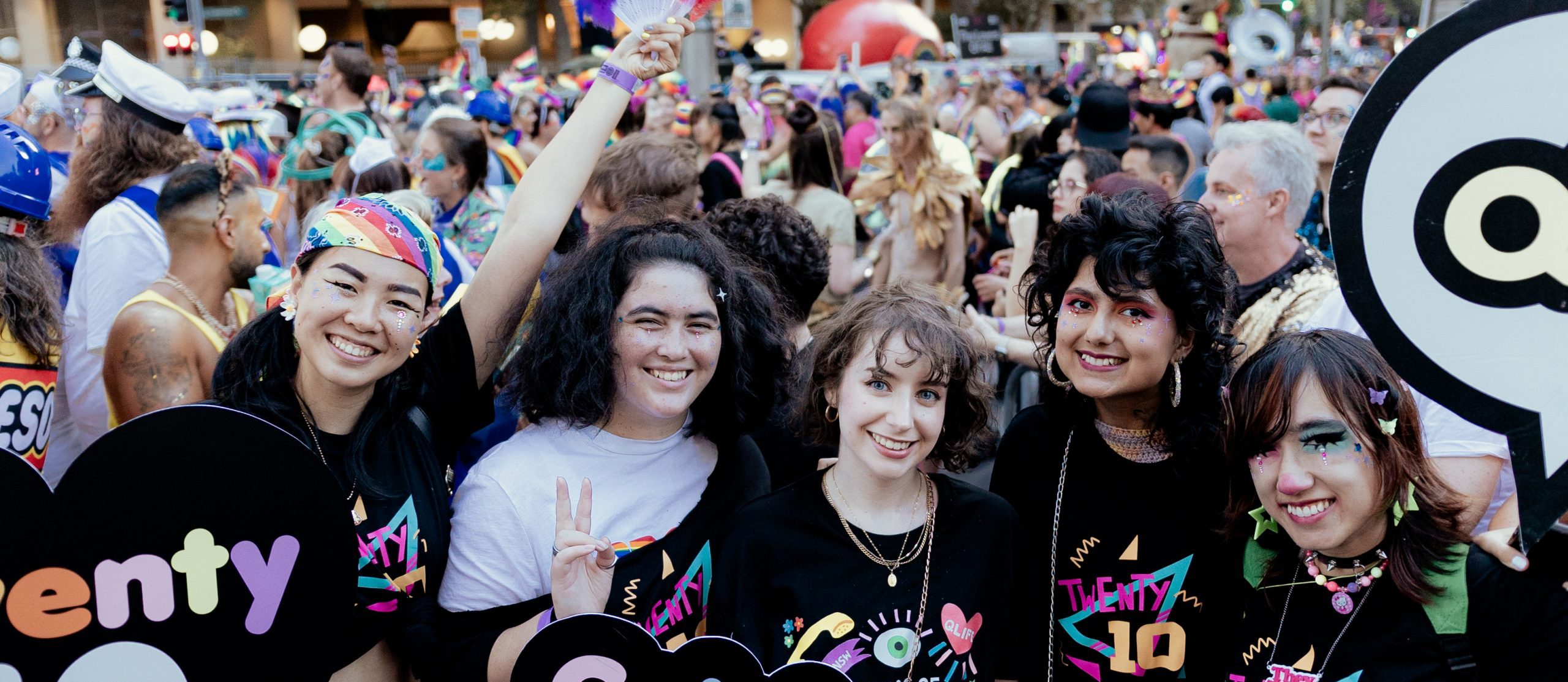 A photo of five young people wearing black tshirts with designs featuring Twenty10 in stylised text. They are dressed up in 80s style makeup and hair.