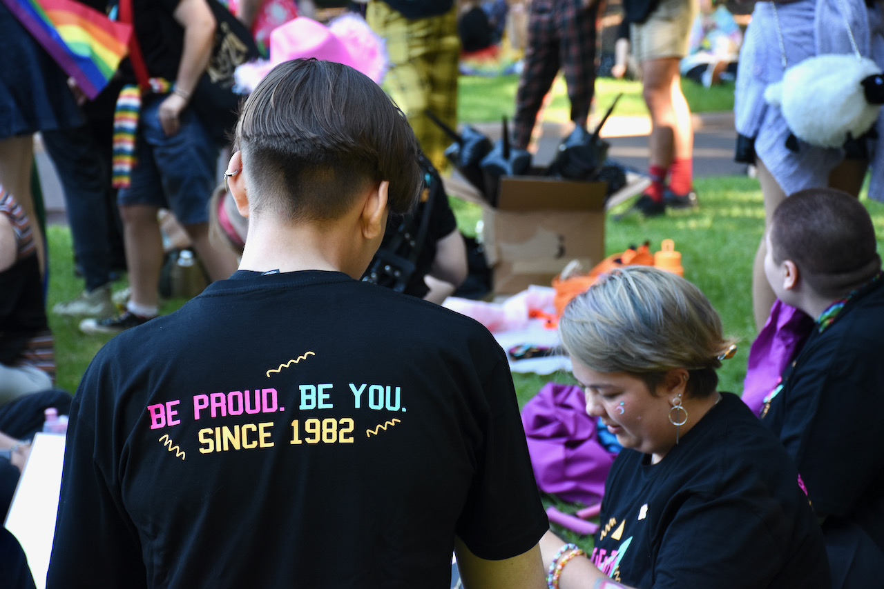 Photo of a young person facing away from the camera. They are wearing a black tee with the words on the back that read: Be Proud. Be You. Since 1982.
