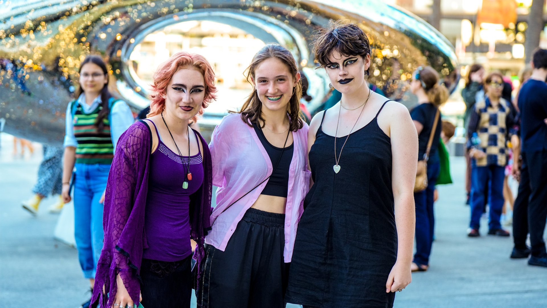 Photo of three young people with dramatic face make up on standing together and looking at the camera. There is a large metal statue in the background behind them. Photo credits to Ash Penin.