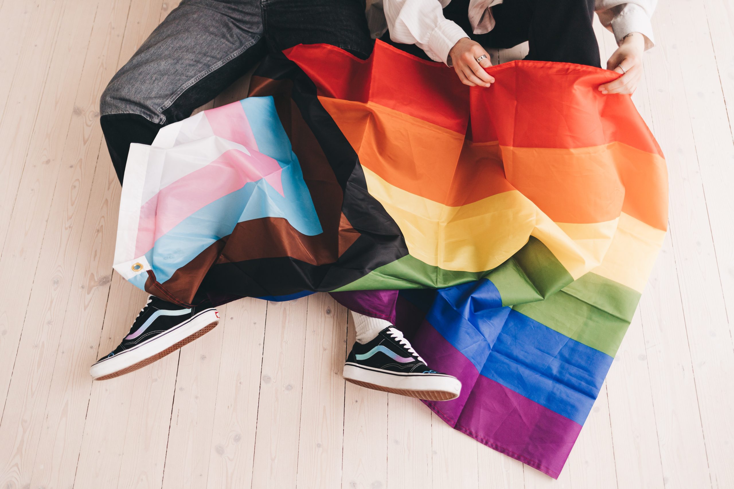 Photo of two people's lower bodies with a progressive Pride flag laid over the top of one person's legs.