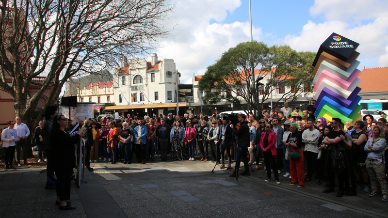 Photo of a crowd of people watching a speaker at the Inner West Pride Square with a large Pride statue in progressive pride colours